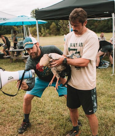 Two men standing together at a race. One man is holding a chicken and the other is holding a megaphone.