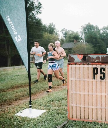 A sign that reads "Psychoactive 2023" next to an Ecotent peak flag in the grass with runners in the background.